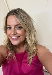Smiling woman with wavy blonde hair wearing a pink top against a light background.